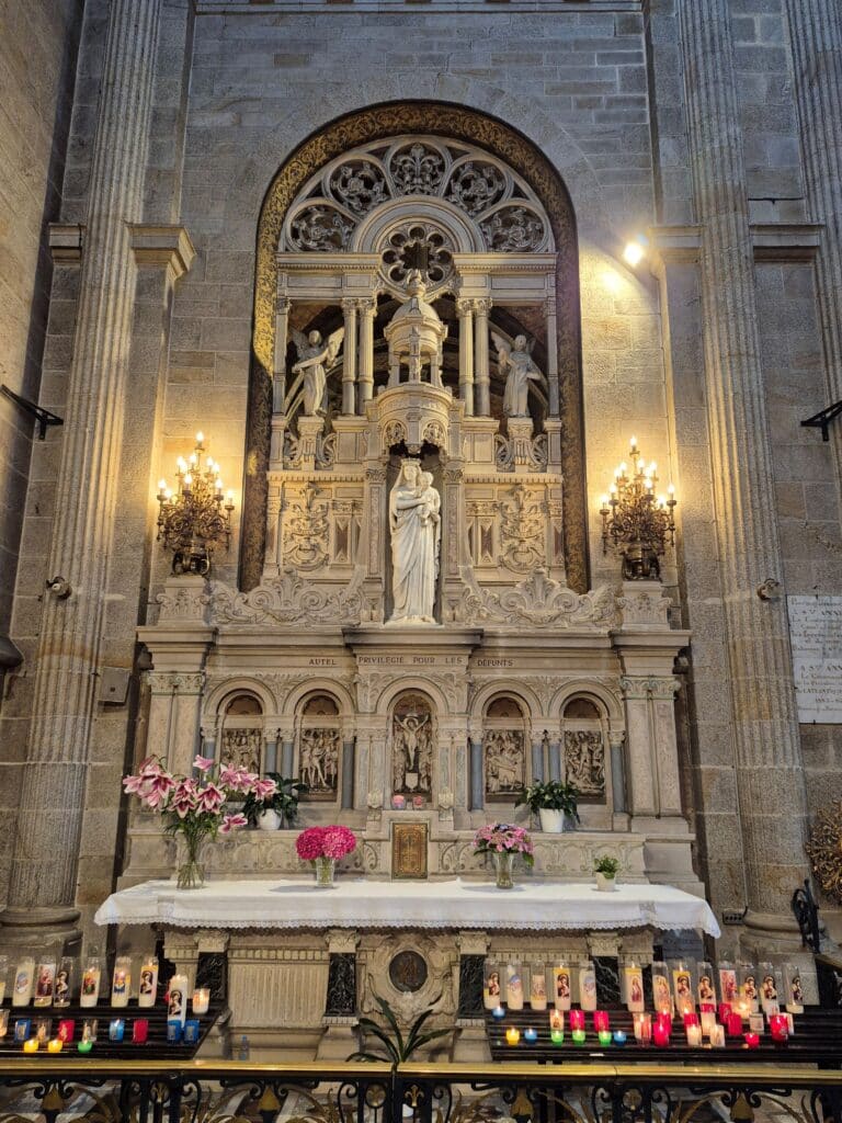 A side altar within the stone Basilica of Sainte-Anne-d’Auray. In the center, a white marble statue of the Virgin Mary holding the Christ Child is framed by an ornate Gothic-style canopy. Below, a white-clad altar is adorned with fresh pink lilies and hydrangeas. Rows of glowing votive candles and prayer intentions in colorful glass holders sit in the foreground.