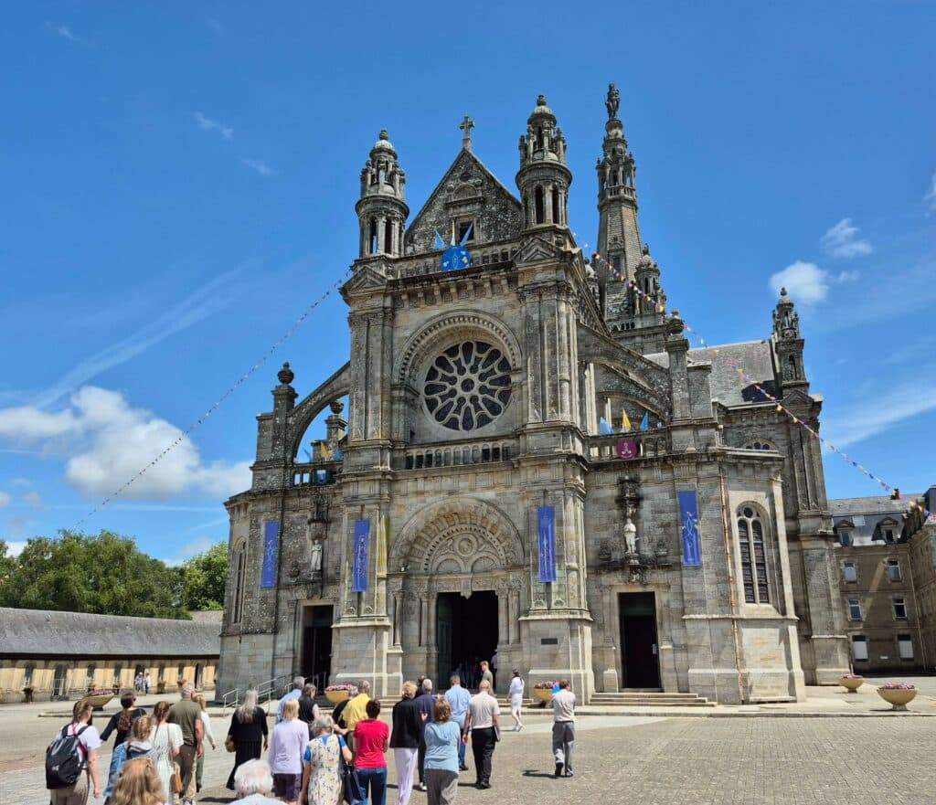 A wide-angle view of pilgrims walking across the stone plaza toward the granite facade and arched entrance of the Basilica of Sainte-Anne-d’Auray in Brittany.