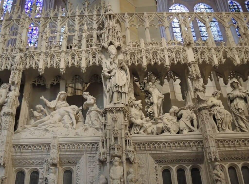 "A side-by-side view of two high-relief stone carvings on the Chartres choir screen. On the left, the Lamentation (Pietà) shows a sorrowful Mary cradling the limp body of Christ, surrounded by figures with expressive, grieving faces. On the right, the Resurrection depicts a triumphant Christ emerging from the tomb with one hand raised in blessing, while Roman soldiers cower in the foreground. Both scenes are framed by the signature 'stone lace'—intricate, deep-cut Gothic canopies that resemble delicate embroidery.