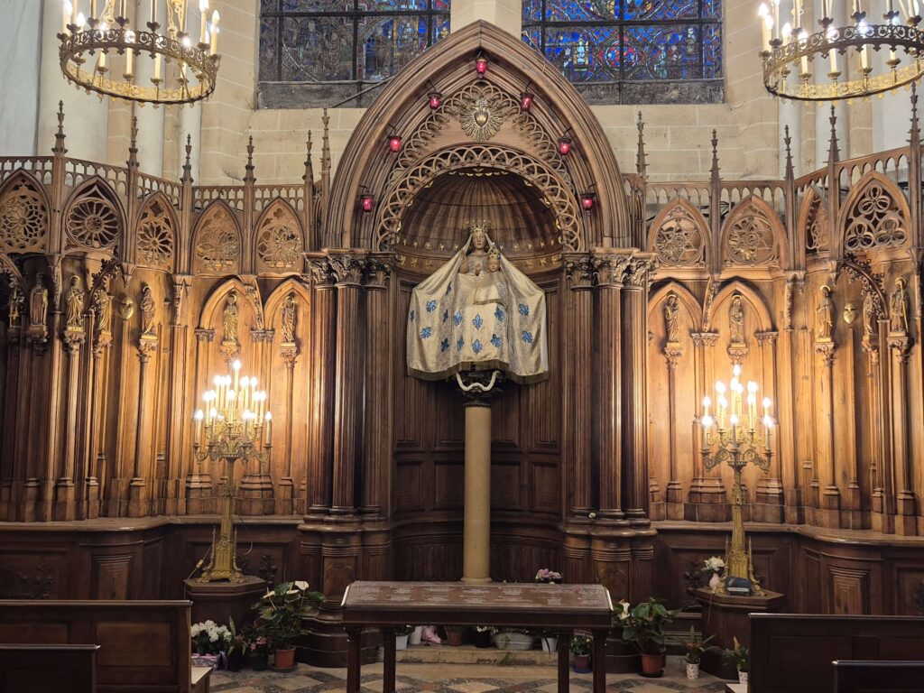 A 16th-century dark pearwood statue of Our Lady of the Pillar at Chartres Cathedral. Mary holds the Christ Child while standing atop a tall stone column, surrounded by candles and ornately carved wood panels