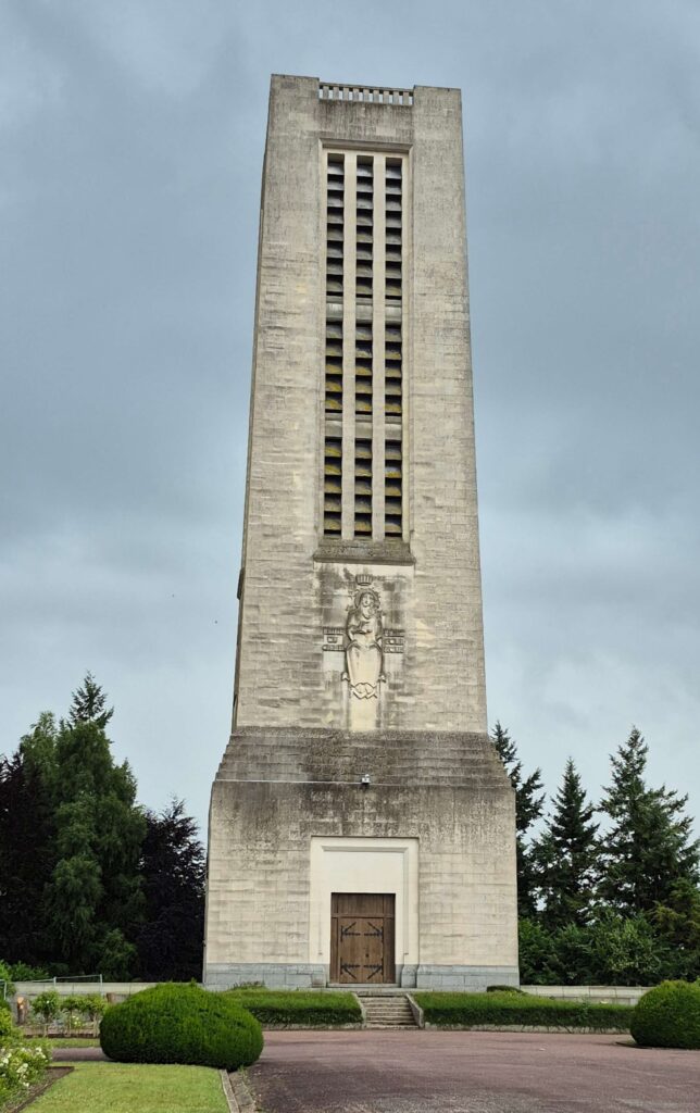 A low-angle view of the large, modern concrete bell tower of the Basilica of Saint Thérèse in Lisieux, featuring its characteristic open-structure design and standing against a clear sky, separate from the main church building.