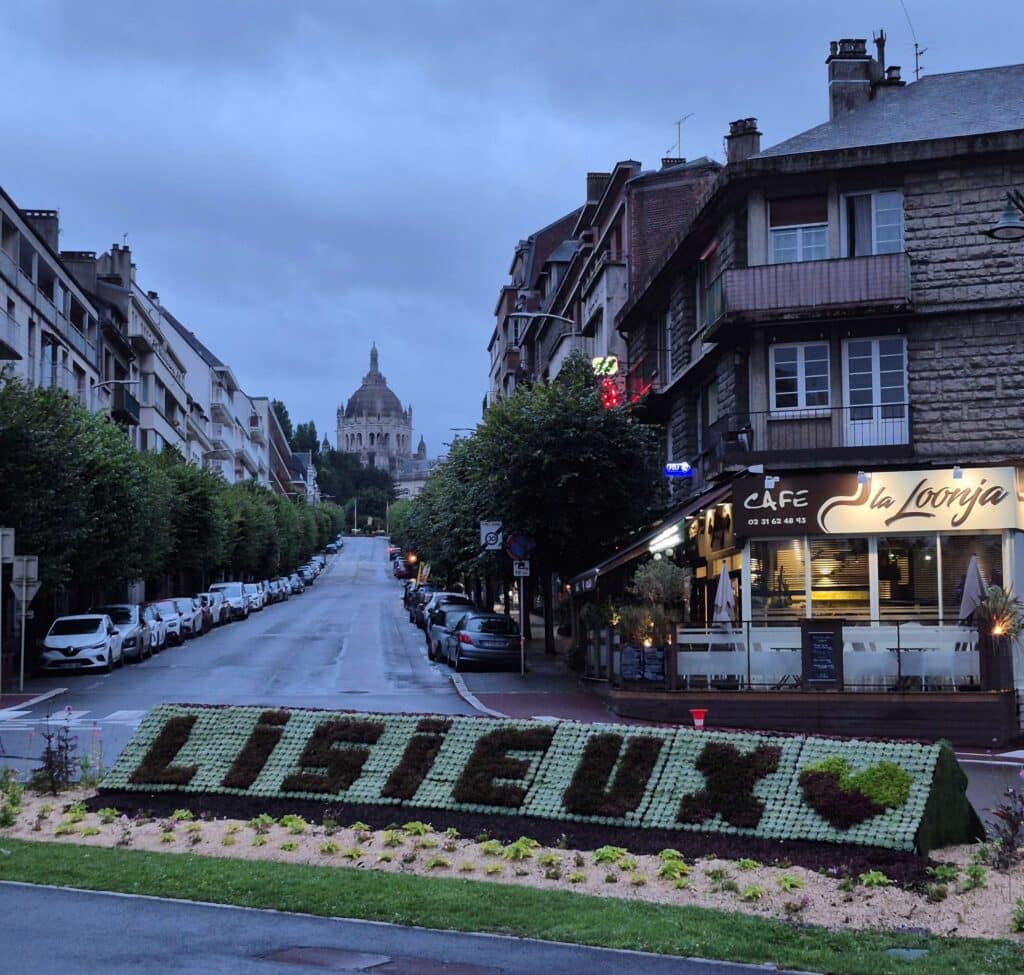 A street-level view looking down a wide boulevard in Lisieux, France, featuring a large public flowerbed in the foreground where the name "LISIEUX" is spelled out with flowers. In the distance, the Basilica of Saint Thérèse is visible, framed by trees and the city streetscape.