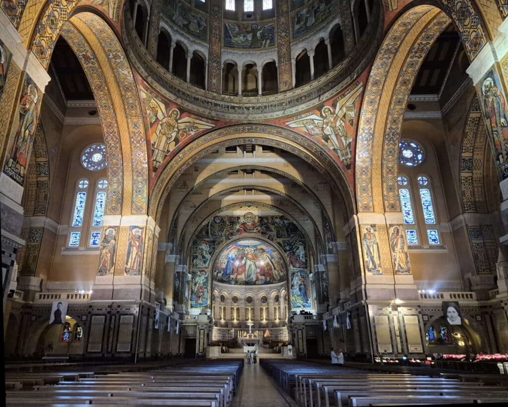 An interior view of the grand nave of the Basilica of Saint Thérèse in Lisieux, featuring high vaulted ceilings covered in intricate golden mosaics, leading the eye toward the ornate high altar and the sunlit apse.