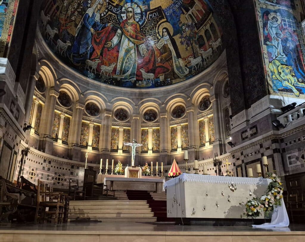 A centered, wide-angle view of the High Altar inside the Basilica of Saint Thérèse of Lisieux, featuring an ornate white marble altar under the mosiac dome flanked by towering mosaic-covered columns and vibrant religious artwork.