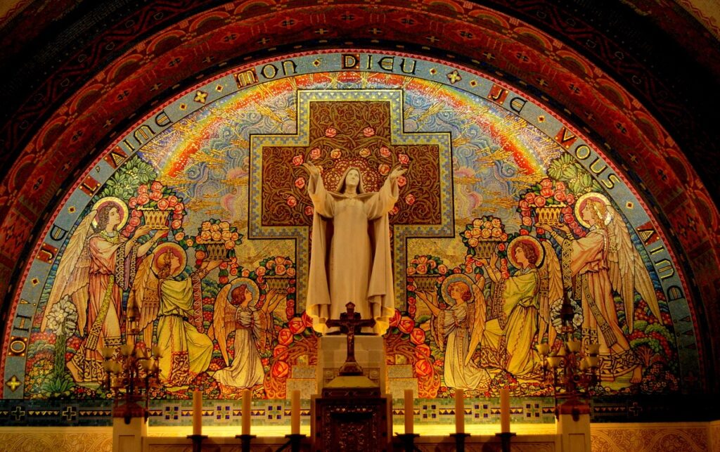 A vibrant, multi-colored mosaic inside the Basilica of St. Thérèse in Lisieux, featuring a central statue of St. Thérèse with uplifted arms surrounded by angels and floral rose motifs against a cross, under an arched alcove.