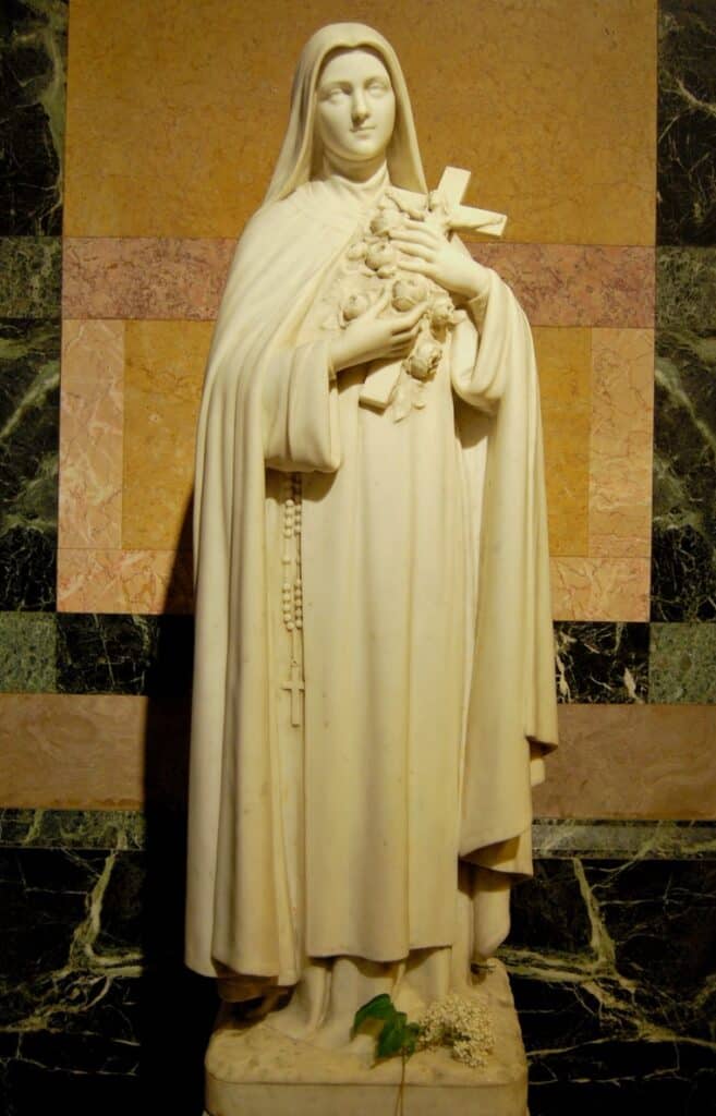 a carved marble statue of Saint Thérèse of Lisieux, depicting her holding a crucifix against her chest, draped in the traditional Carmelite habit, set against the backdrop of the Basilica’s golden interior.